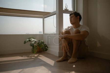 Trauriger und einsamer asiatischer Mann sitzt auf dem Boden und umarmt sein Knie.