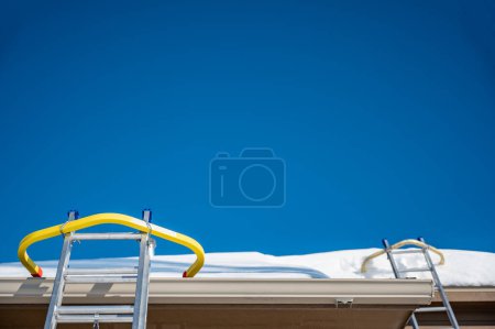 Foto de Ladder and safety hook on a residential roof covered in snow as gutters are being installed. . High quality photo - Imagen libre de derechos