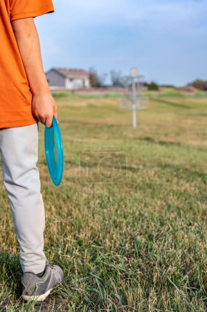 Téléchargez les photos : L'herbe sélective de fairway de foyer avec un jeune enfant déconcentré tenant un disque de putter et un but de golf au loin. . Photo de haute qualité - en image libre de droit