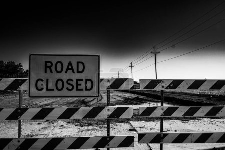 A black and white photograph of a road closed sign standing on the side of an empty street. The sign is prominently displayed, indicating that the road is not accessible to vehicles or pedestrians.