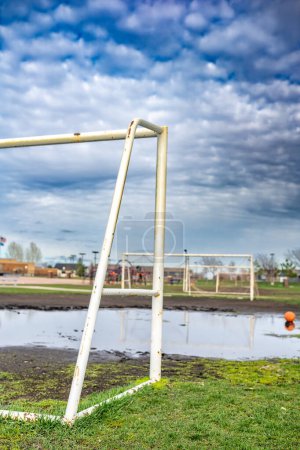 A soccer goal stands prominently in the center of a muddy field, surrounded by splattered soil and grass. The scene suggests recent gameplay or preparation for a match, with the goalpost serving as