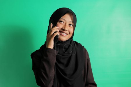 Photo for Cheerful young Asian girl in black hijab or headscarf talking on Cellphone looking aside isolated on green background. - Royalty Free Image