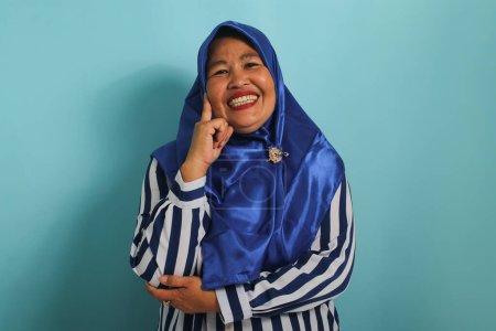 Photo for An excited middle-aged Asian woman, wearing a blue hijab and a striped shirt, gets an amazing idea while standing against a blue background. - Royalty Free Image
