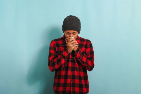 Foto de Un joven asiático llorando con un gorro de gorro y una camisa de franela a cuadros roja limpia las lágrimas con un pañuelo mientras está de pie sobre un fondo azul - Imagen libre de derechos