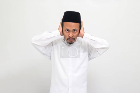 Téléchargez les photos : Un jeune Indonésien en colère portant une chanson, un peci ou une kopiah, se couvrant les oreilles, se sentant mécontent, contrarié et déçu, isolé sur un fond blanc - en image libre de droit