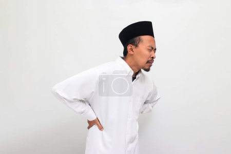 Téléchargez les photos : Jeune Indonésien portant une chanson ou peci ou kopiah, souffre d'un mal de dos, éprouvant des maux de dos, isolé sur un fond blanc - en image libre de droit