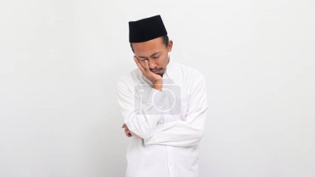 Téléchargez les photos : Un jeune Indonésien drôle portant une chanson ou peci ou kopiah, garde sa main sur ses joues, l'air ennuyé, isolé sur un fond blanc - en image libre de droit