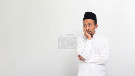 Téléchargez les photos : Jeune Indonésien ennuyé portant une chanson ou peci ou kopiah, garder la main sur le menton tout en regardant de côté l'espace de copie vide, isolé sur un fond blanc - en image libre de droit