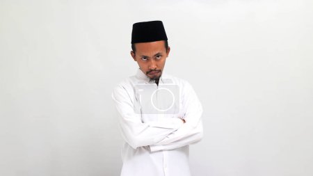 Téléchargez les photos : Jeune Indonésien ennuyé portant une chanson ou peci ou kopiah, garde son bras plié en regardant la caméra, isolé sur un fond blanc - en image libre de droit