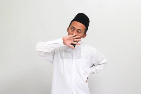 Téléchargez les photos : Un jeune Indonésien fatigué drôle portant une chanson ou peci ou kopiah, bâille, couvrant sa bouche avec sa main, se sentant épuisé par le surmenage et l'insomnie, isolé sur un fond blanc - en image libre de droit