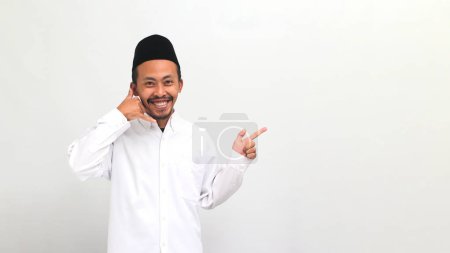 Téléchargez les photos : Jeune Indonésien excité portant une chanson ou peci ou kopiah, fait un geste de m'appeler tout en pointant de côté à l'espace de copie vide, isolé sur un fond blanc - en image libre de droit