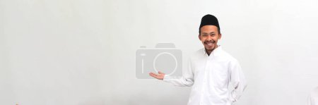 Téléchargez les photos : Jeune Indonésien excité, portant une chanson, peci, ou kopiah, étend ses mains sur le côté de l'espace de copie, invitant quelqu'un à venir, isolé sur fond blanc - en image libre de droit