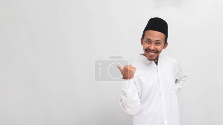 Téléchargez les photos : Jeune Indonésien, portant une chanson, peci, ou kopiah, pointe le pouce de côté à l'espace de copie vide, isolé sur fond blanc - en image libre de droit