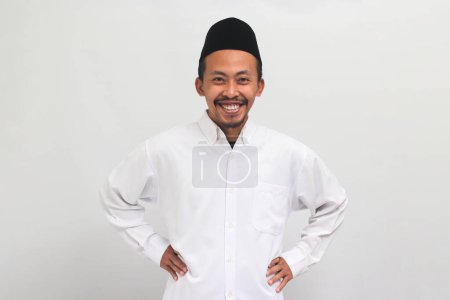 Téléchargez les photos : Un jeune Indonésien confiant portant une chanson, un peci ou une kopiah, garde la main sur sa taille dans un geste akimbo, isolé sur un fond blanc - en image libre de droit