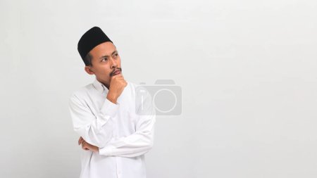 Téléchargez les photos : Le jeune Indonésien confus, portant une chanson, un peci ou une kopiah, croise les bras avec la main sur le menton, réfléchissant pensivement en regardant l'espace vide, isolé sur un fond blanc - en image libre de droit