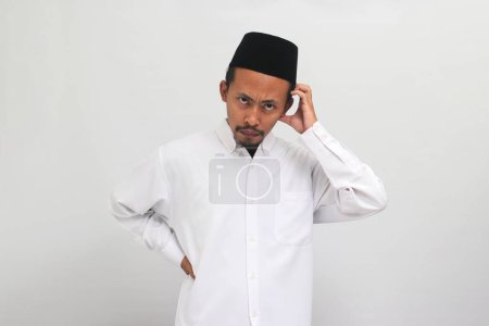 Téléchargez les photos : Jeune Indonésien confus, portant une chanson, peci, ou kopiah, se gratte la tête en regardant la caméra, isolé sur un fond blanc - en image libre de droit