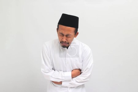 Téléchargez les photos : Un jeune Indonésien malheureux, portant une chanson, un peci ou une kopiah, souffre d'une douleur à l'estomac, se penchant et tenant ses mains sur son ventre, isolé sur un fond blanc - en image libre de droit