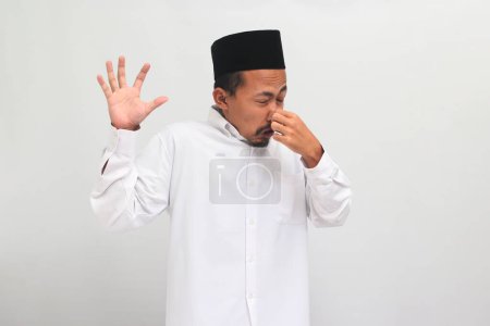 Téléchargez les photos : Homme indonésien agacé, portant une chanson, peci, ou kopiah, retient son souffle, avec ses doigts sur le nez, comme il sent quelque chose puant, dégoûtant, et intolérable, isolé sur un fond blanc - en image libre de droit