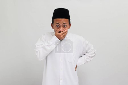 Téléchargez les photos : Embarrassé Jeune Indonésien, portant une chanson, peci, ou kopiah, est choqué, couvrant sa bouche en réponse à une erreur et se sentant mal pour dire quelque chose de mal, isolé sur un fond blanc - en image libre de droit