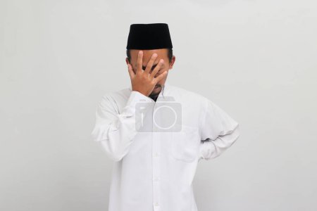 Téléchargez les photos : Un jeune Indonésien honteux et embarrassé, portant une chanson, un peci ou une kopiah, des facepalms, couvrant son visage de sa main, isolé sur un fond blanc - en image libre de droit