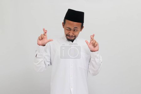 Téléchargez les photos : Jeune Indonésien excité, portant une chanson, peci, ou kopiah, fait un geste croisé des doigts et regarde de côté l'espace de copie vide, isolé sur un fond blanc - en image libre de droit