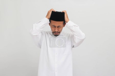 Téléchargez les photos : Jeune Indonésien, portant une chanson, peci, ou kopiah, souffre d'un mal de tête, se sentant désespéré et stressé en raison de la douleur et de la migraine, isolé sur fond blanc - en image libre de droit