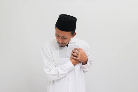 Téléchargez les photos : Jeune Indonésien, portant une chanson, peci, ou kopiah, saisit sa poitrine, éprouvant une douleur aiguë, qui peut être un symptôme d'une crise cardiaque, isolé sur fond blanc - en image libre de droit