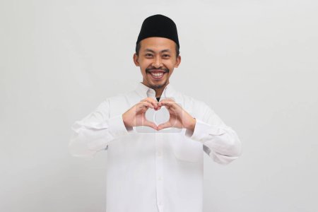 Téléchargez les photos : Jeune Indonésien souriant, portant une chanson, peci, ou kopiah, montre un geste d'amour avec sa main, isolé sur fond blanc - en image libre de droit