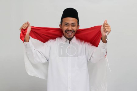 Téléchargez les photos : Un jeune Indonésien excité, portant une chanson, un peci ou une kopiah, tient un drapeau indonésien pour célébrer la fête de l'indépendance de l'Indonésie le 17 août, isolé sur fond blanc - en image libre de droit