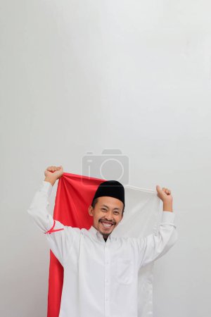 Téléchargez les photos : Un jeune Indonésien excité, portant une chanson, un peci ou une kopiah, tient un drapeau indonésien pour célébrer la fête de l'indépendance de l'Indonésie le 17 août, isolé sur fond blanc - en image libre de droit