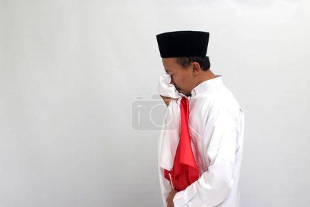 Téléchargez les photos : Jeune Indonésien, portant une chanson, peci, ou kopiah, embrasse le drapeau indonésien comme un symbole de respect tout en célébrant la fête de l'indépendance de l'Indonésie le 17 août, isolé sur fond blanc - en image libre de droit