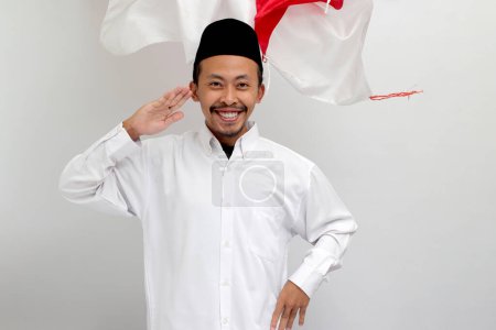 Téléchargez les photos : Le jeune Indonésien excité, vêtu d'une chanson, d'un peci ou d'une kopiah, fait un geste de salut avec le drapeau indonésien en arrière-plan tout en célébrant le Jour de l'indépendance de l'Indonésie le 17 août - en image libre de droit