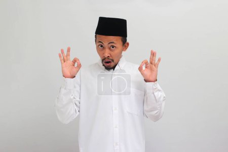 Téléchargez les photos : Surpris jeune Indonésien, portant une chanson, peci, ou kopiah, regarde la caméra tout en faisant le signe OK geste, isolé sur fond blanc - en image libre de droit