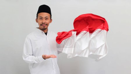 Téléchargez les photos : Un jeune Indonésien portant une chanson, un peci ou une kopiah arbore fièrement le drapeau indonésien à ses côtés, isolé sur fond blanc. - en image libre de droit