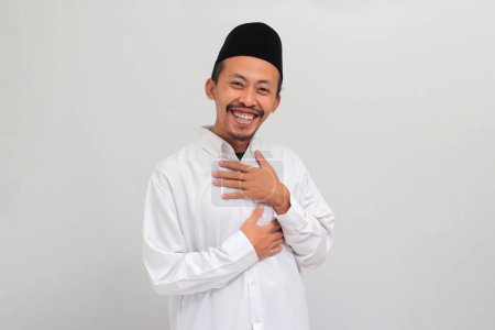 Téléchargez les photos : Un jeune indonésien reconnaissant, portant une chanson, peci, ou kopiah, tient ses mains sur son cœur, se sentant soulagé et reconnaissant, isolé sur fond blanc - en image libre de droit