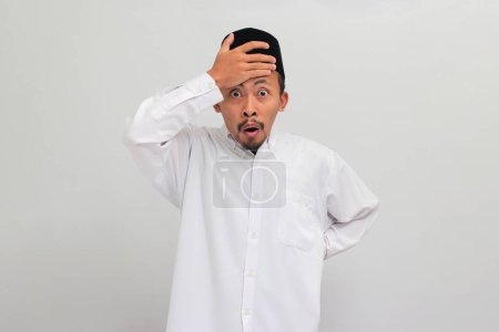 Téléchargez les photos : Choqué et inquiet Jeune Indonésien, portant une chanson, peci, ou kopiah, tenir la main sur la tête, la bouche ouverte et l'air effrayé, oublier se rappeler quelque chose de mauvais, préoccupé, isolé sur fond blanc - en image libre de droit