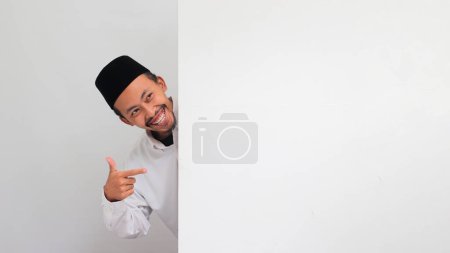 Téléchargez les photos : Un jeune Indonésien excité, portant une chanson, un peci ou une kopiah, se tient derrière une bannière blanche vierge et pointe un espace de copie vide sur un panneau publicitaire, isolé sur fond blanc - en image libre de droit