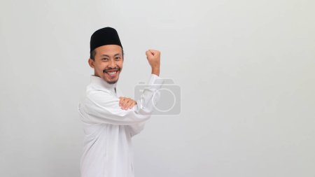 Téléchargez les photos : Jeune Indonésien confiant, portant une chanson, un peci ou une kopiah, faisant un geste fort, montrant de la force en fléchissant ses bras et ses muscles vers la caméra, isolé sur fond blanc - en image libre de droit