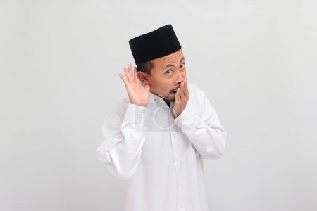Téléchargez les photos : Un jeune Indonésien excité, portant une chanson, un peci ou une kopiah, sourit en plaçant sa main sur son oreille, écoutant des rumeurs ou des commérages, isolé sur fond blanc - en image libre de droit