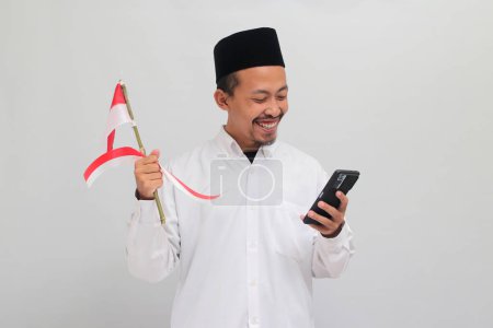 Téléchargez les photos : Le jeune Asiatique excité, vêtu d'une chanson, d'un peci ou d'une kopiah, célèbre le jour de l'indépendance de l'Indonésie le 17 août alors qu'il tient le drapeau indonésien et un téléphone portable, isolé sur fond blanc - en image libre de droit