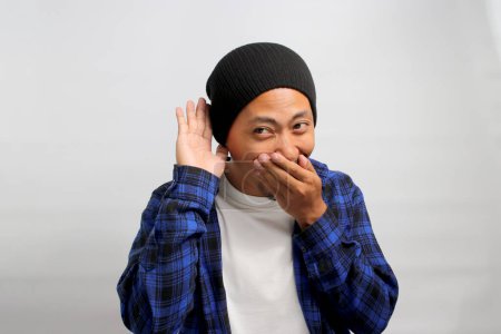 Foto de Joven hombre asiático, vestido con un sombrero de gorro y camisa casual, se ríe mientras escucha atentamente, la mano tapó su oreja, posiblemente atrapando el viento de rumores o chismes mientras está de pie contra el fondo blanco - Imagen libre de derechos