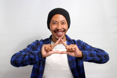 Téléchargez les photos : Jeune homme asiatique, vêtu d'une chemise décontractée et coiffé d'un bonnet, crée une forme de triangle avec ses mains, symbolisant le concept de protection de l'environnement, de recyclage et de réutilisation - en image libre de droit