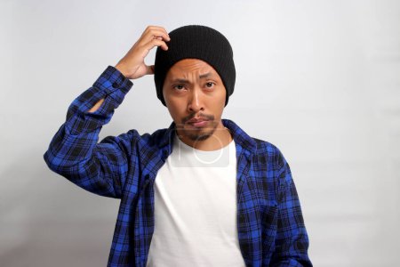 Photo for A puzzled young Asian man, dressed in a beanie hat and casual clothes, scratches his head while seeking a solution, portraying doubt and confusion, standing against a white background - Royalty Free Image