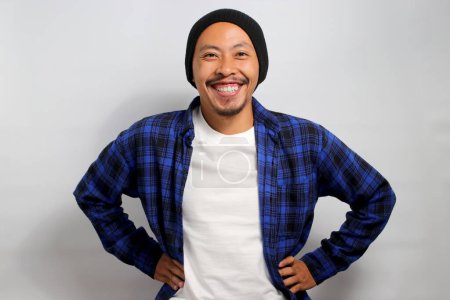 Photo for Optimistic young Asian man, dressed in a beanie hat and casual shirt, stands confidently with arms akimbo, smiling at the camera, exuding confidence. Standing against white background. - Royalty Free Image