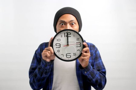 Photo for An Asian man, sporting a beanie hat and casual shirt, holds a clock in front of his face, looking at the camera with a surprised and humorous expression, while standing against a white background - Royalty Free Image