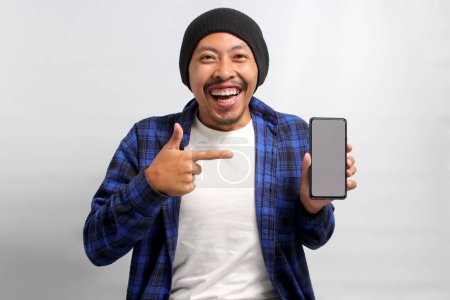 Photo for Excited Asian man, dressed in a beanie hat and casual shirt, excitedly showcases and points towards a mobile phone with an empty white screen, suggesting or recommending a new app or a special offer - Royalty Free Image