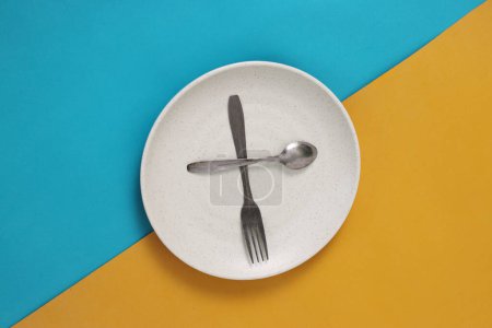 Empty plate with spoon and fork on a blue and yellow background, representing fasting during Ramadan and the anticipation of breaking fast