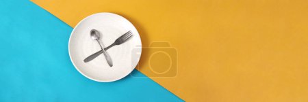 Empty plate with spoon and fork on a blue and yellow background with empty copy space for text or advertisement, representing fasting during Ramadan and the anticipation of breaking fast