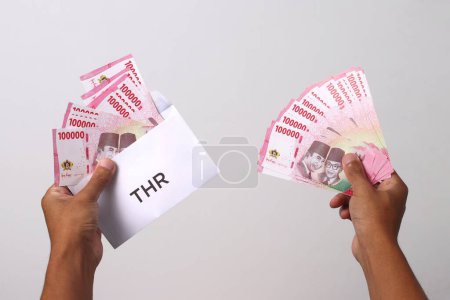 Hand in Hand einen Umschlag mit indonesischen Rupiah-Banknoten. THR oder Tunjangan Hari Raya ist ein Urlaubsgeld oder Bonus, der traditionell während des Ramadan an Angestellte und Bedürftige in der Nähe vergeben wird.