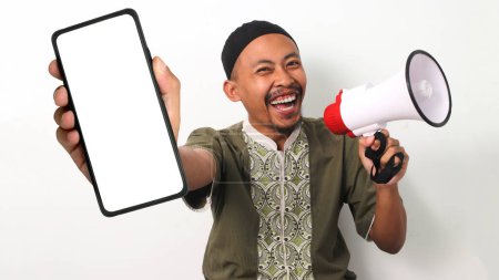 Un homme musulman indonésien excité en koko et en peci montre un écran de téléphone vierge et tient un mégaphone, faisant la promotion d'une nouvelle application ou d'une vente Ramadan. Isolé sur fond blanc
